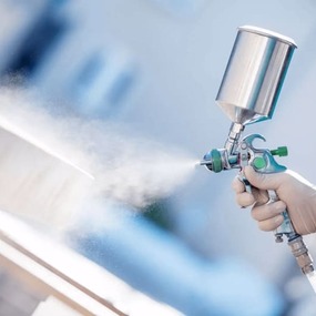 Eine Hand sprüht mit einer Lackierpistole Farbe auf eine Holzoberfläche im Freien.